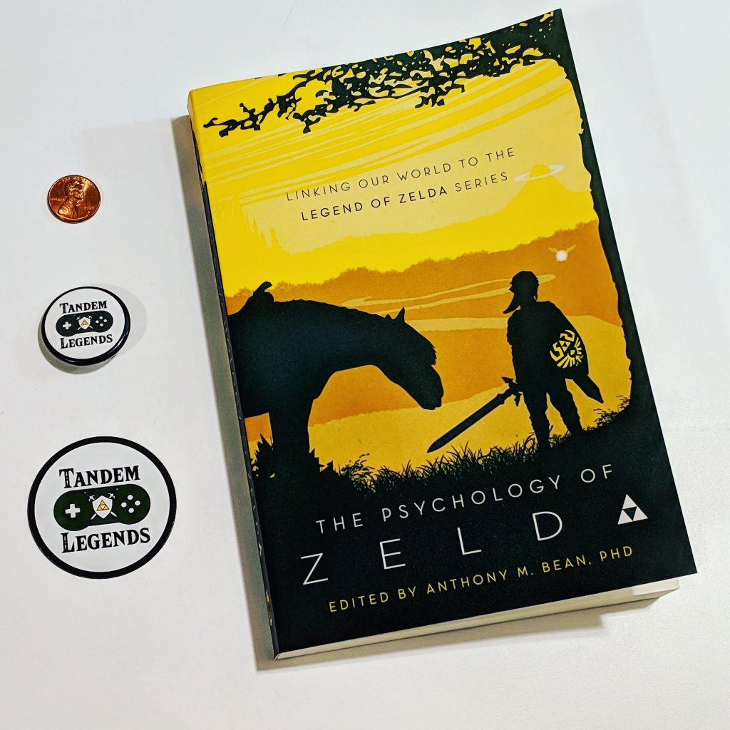 Cover of Psychology of Zelda, a Tandem Legends branded Pin, a Tandem Legends branded magnet, and a penny for scale